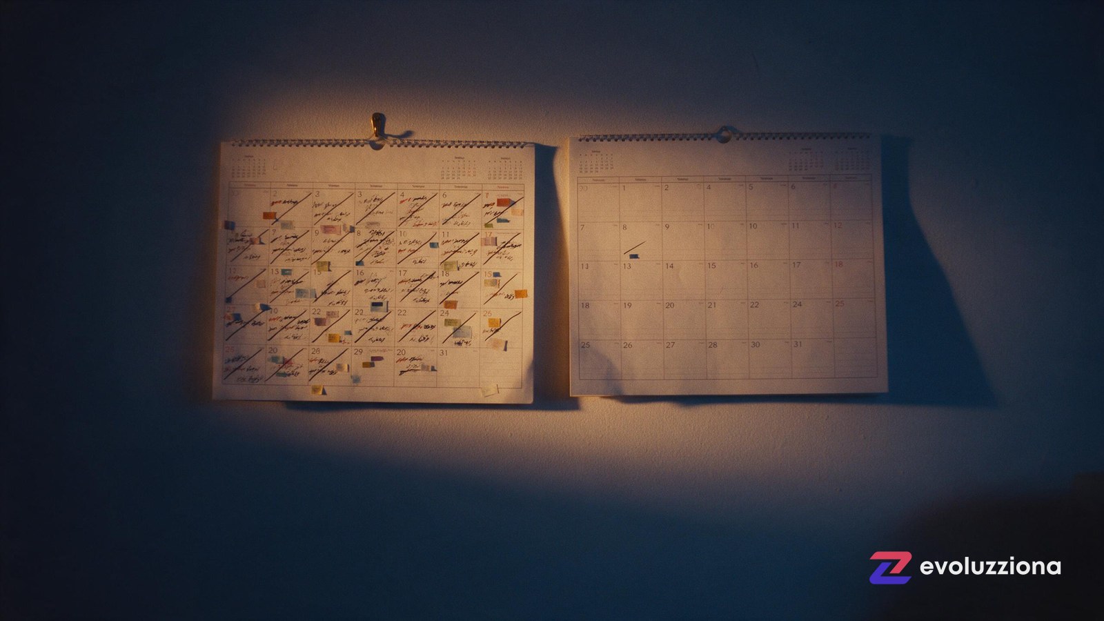 Dos calendarios de pared colgados juntos, uno lleno de marcas ordenadas y otro casi vacío, con luz lateral cálida