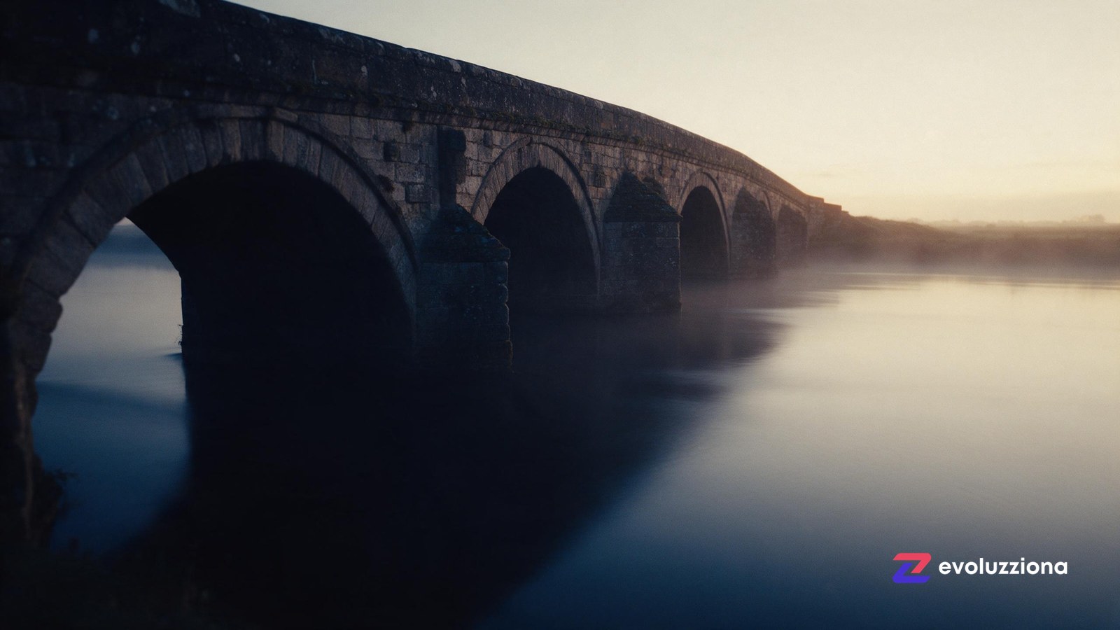 Puente de piedra al amanecer sobre agua en calma, con luz cálida en el extremo lejano